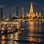 A public river ferry passing the illuminated Bangkok skyline after dark