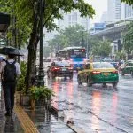 People walking with umbrellas beside Bangkok traffic during a normal tropical rain shower