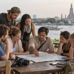 Six adult travellers reviewing a Thailand route with a local coordinator beside the Chao Phraya River in Bangkok